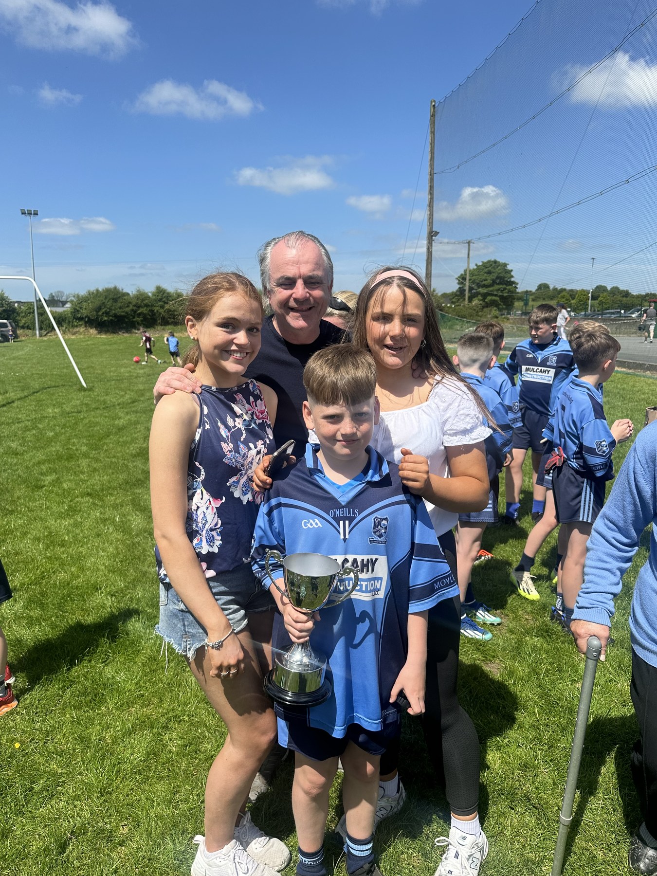 A group on a sunny sports field with a young player holding a trophy.