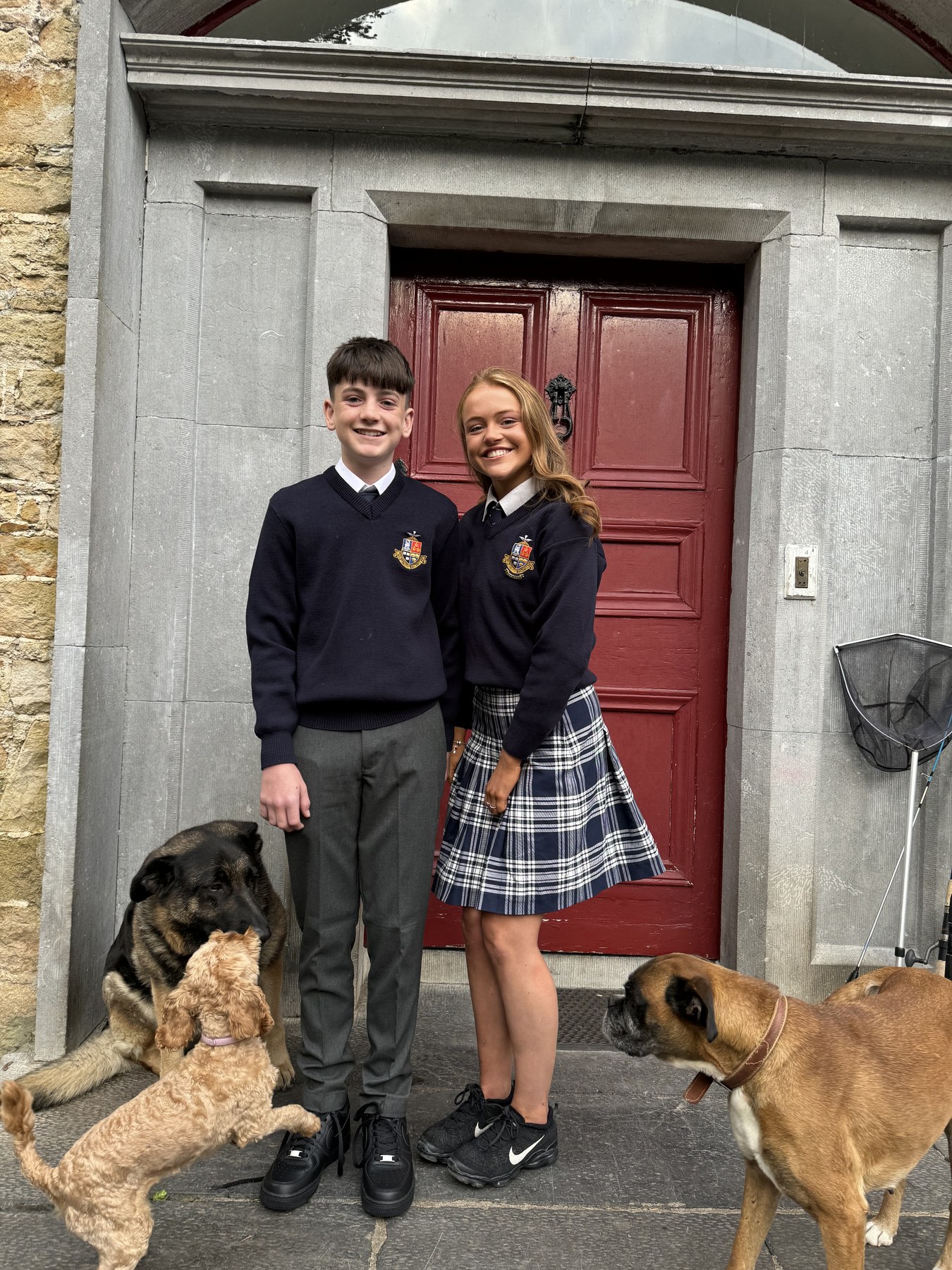 Two students in school uniforms standing by a red door with two dogs.